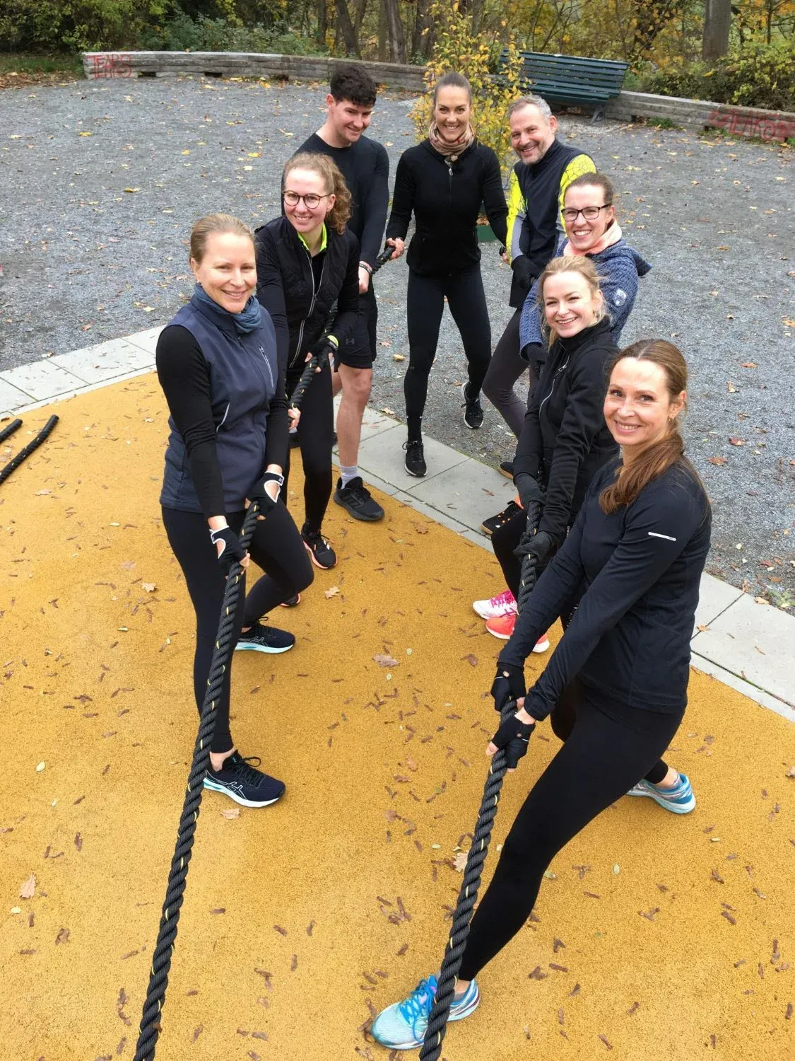 Trainingsgruppe mit Battle Ropes auf einem Outdoor-Sportplatz im Herbst, alle lächeln in die Kamera