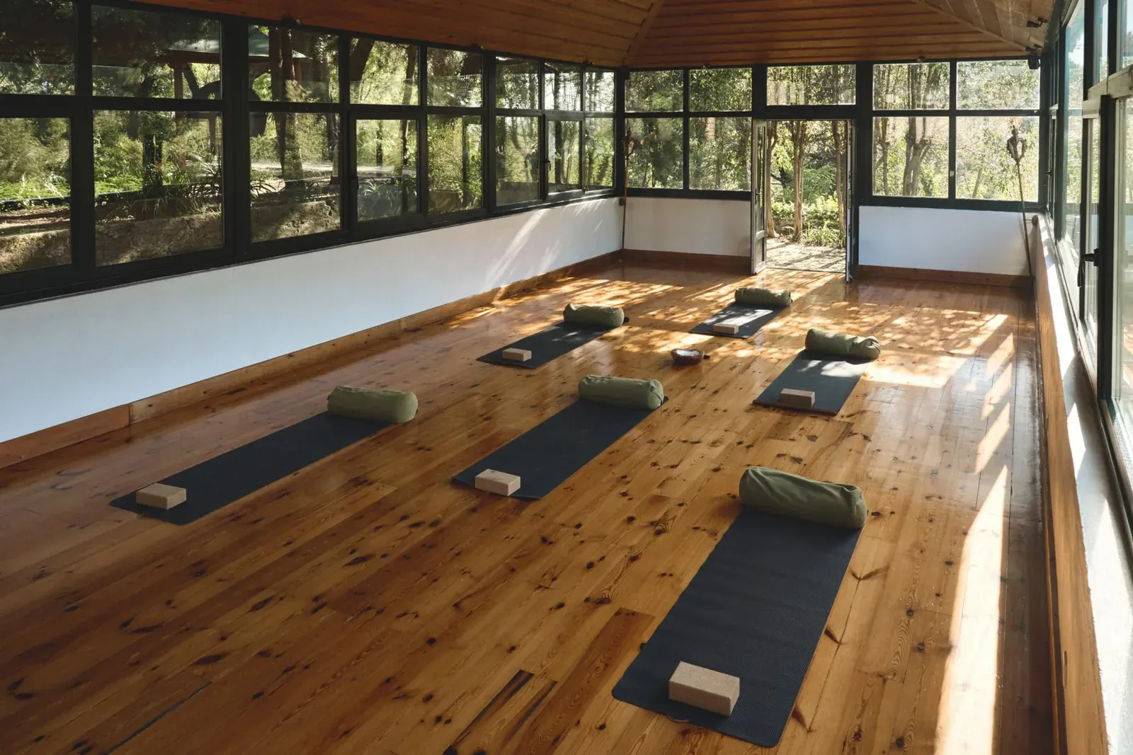 Heller Retreatraum mit Fensterfront zum Wald, Yogamatten, Kissen und Blöcken auf dem Holzboden